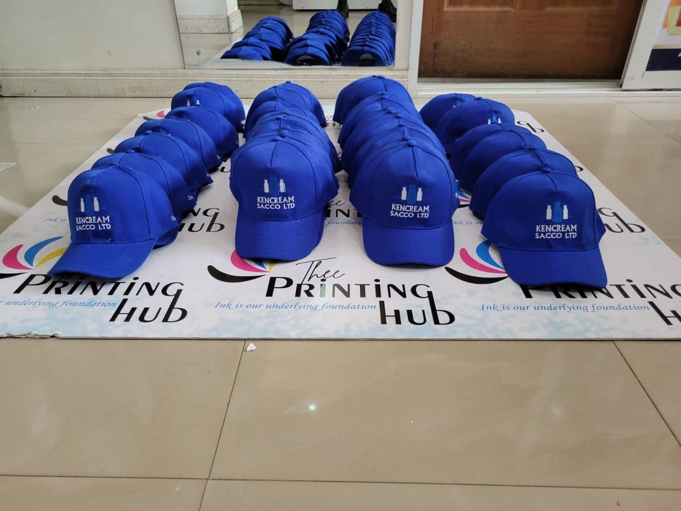 Rows of blue branded caps laid out on a Thee Printing Hub banner